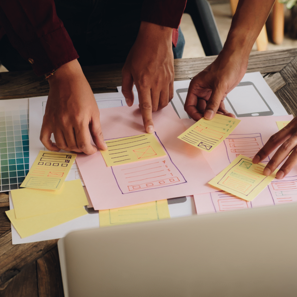 Hands collaborating on digital marketing strategy, arranging colorful sticky notes and mockups on a workspace, emphasizing teamwork and planning for effective online growth.